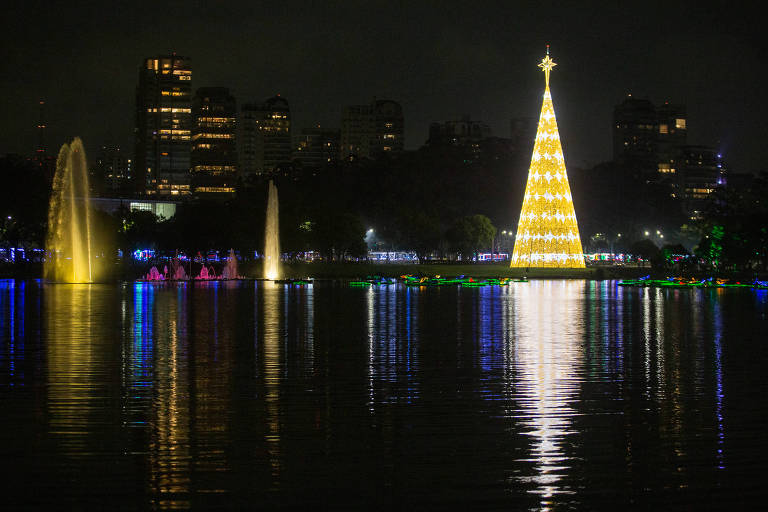 imagem geral mostra árvore de natal de são paulo no lago do parque ibirapuera, com prédios ao fundo; luzes são refletidas na água, da árvore de natal a direita e das fontes coloridas a esquerda 