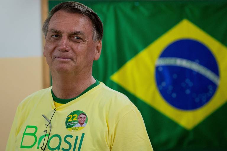 Jair Bolsonaro é homem branco, está sorrindo para a foto. Veste camiseta amarela com gola verde e Brasil escrito em verde. Ao fundo, bandeira do Brasil