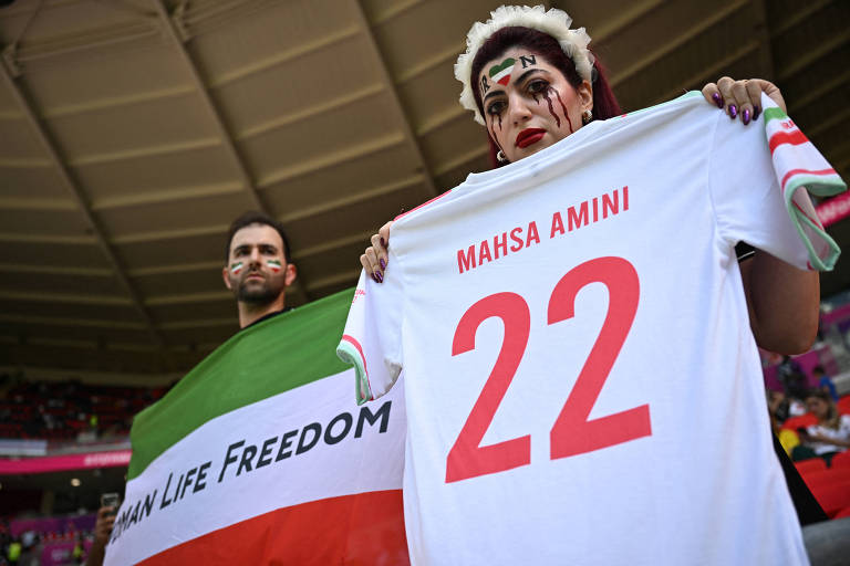 Mulher segura camiseta do Irã com o nome de Mahsa Amini e homem segura bandeira com as cores do país e as palavras Women Life Freedom