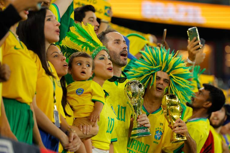 Torcedores do Brasil seguram troféus da copa do mundo simulados enquanto cantam seu hino nacional antes do início da partida entre Brasil e Coreia do Sul no Estádio 974 em Doha pelas oitavas de final