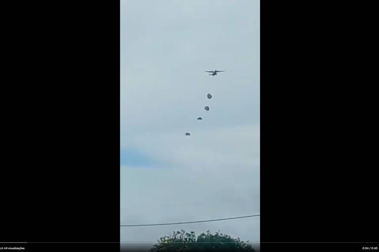 Captura de frame de vídeo mostra céu azul com nuvens ralas e avião bem no alto, pequeno na imagem, com outras imagens menores enfileiradas, que são ps paraquedistas. Mais para baixo há um fio de poste e topo de copa