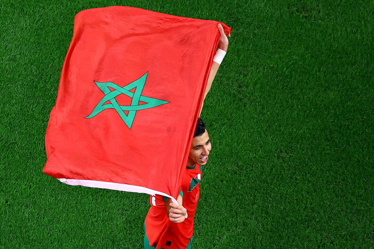 O zagueiro Jawad El Yamiq estende sobre a cabeça a bandeira de Marrocos no estádio Cidade da Educação depois da classificação da seleção para seu segundo mata-mata no Mundial no Qatar; a bandeira é vermelha e tem uma estrela de cinco pontas verde no centro