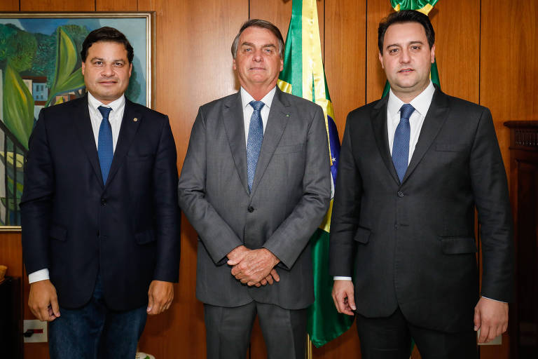 foto de três homens de terno com bandeira brasileira ao fundo em gabinete de paredes de madeira