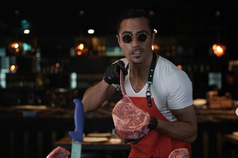 Em foto colorida, homem de blusa branca e avental vermelho separa as carnes para preparar um prato