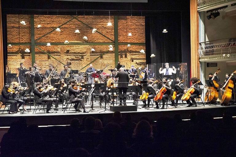 Em foto colorida, a Orquestra Tom Jobim aparece tocando em um palco