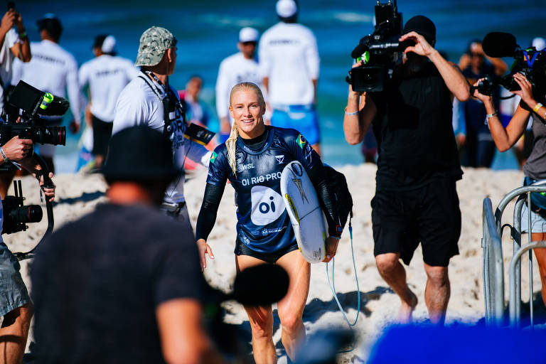 A surfista Tatiana Weston-Webb caminha pela praia segurando sua prancha e com muitos fotógrafos ao redor dela