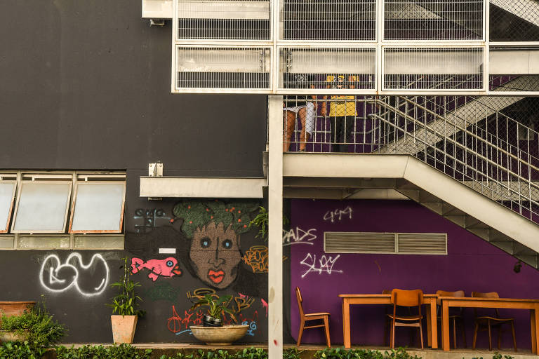 Fachada da escola, com grafites coloridos em uma parede pintada de roxo, algumas pichações e uma escada externa ao prédio