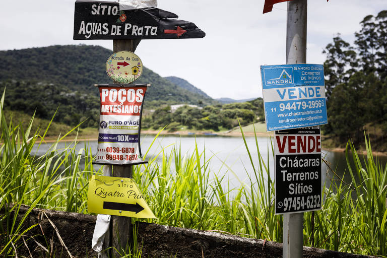 Placas de compra, venda e regularização de terrenos em estrada. Ao fundo, é possível ver vegetação e parte da represa de Atibainha