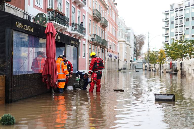 Imagem mostra uma rua larga, com prédios baixos e coloridos, inundada pelas águas da chuva. Em destaque um grupo de bombeiros que trabalha para acelerar a drenagem