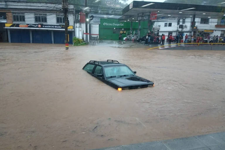 Imagem mostra um carro preto submerso em rua inundada
