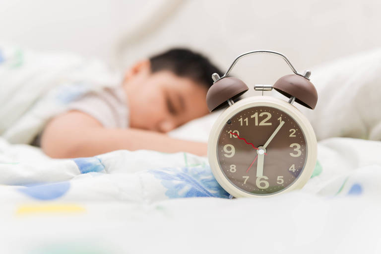 À frente, um relógio do tipo despertador; ao fundo, uma criança dorme
