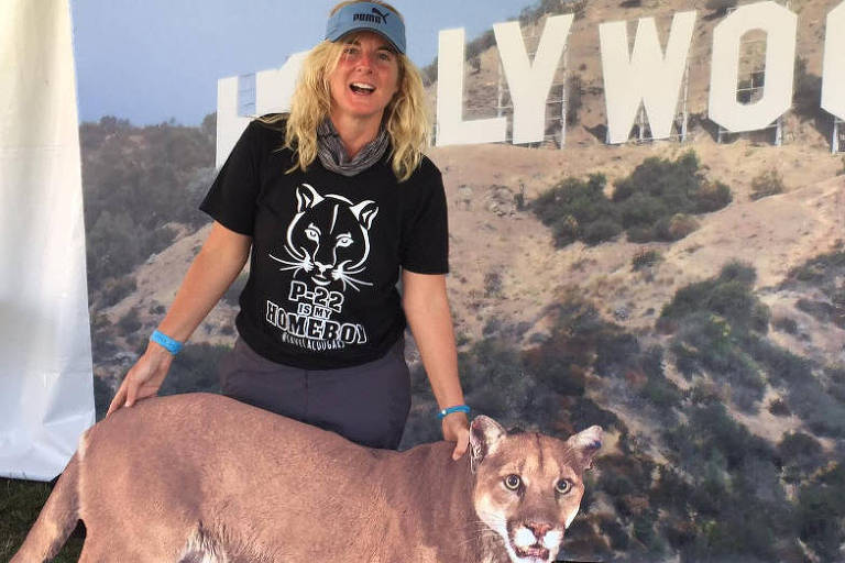 Mulher loira segura cartaz de animal; ao fundo há cartaz com o letreiro de Hollywood