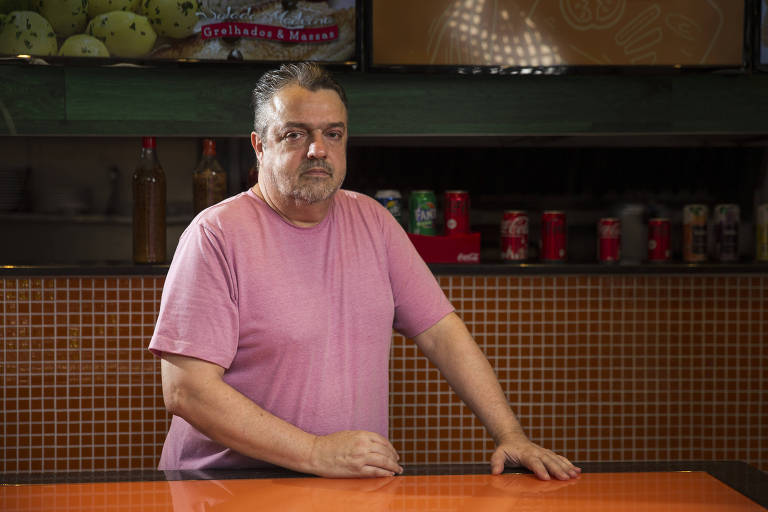 Retrato de Edgar, um homem branco, com cabelos e barba curtos e grisalhos; ele veste camiseta rosa e está do balcão de um restaurante