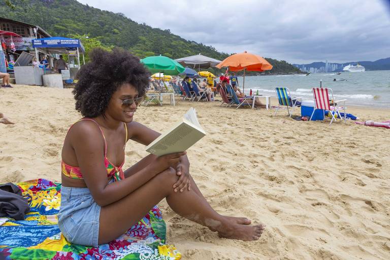 mulher negra lê em praia com cadeiras e guarda-sois ao fundo