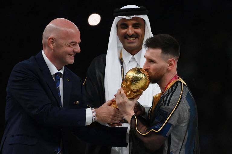 No estádio Lusail, o presidente da Fifa, Gianni Infantino, segura a Taça Fifa enquanto Messi dá um beijo no troféu, observados pelo xeque Tamim bin Hamad al-Thani