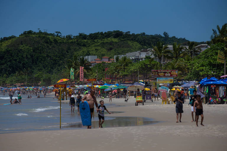 A imagem mostra uma praia movimentada com várias pessoas caminhando na areia e na água. Ao fundo， há barracas coloridas e vegetação nas colinas. O céu está limpo e azul， indicando um dia ensolarado.