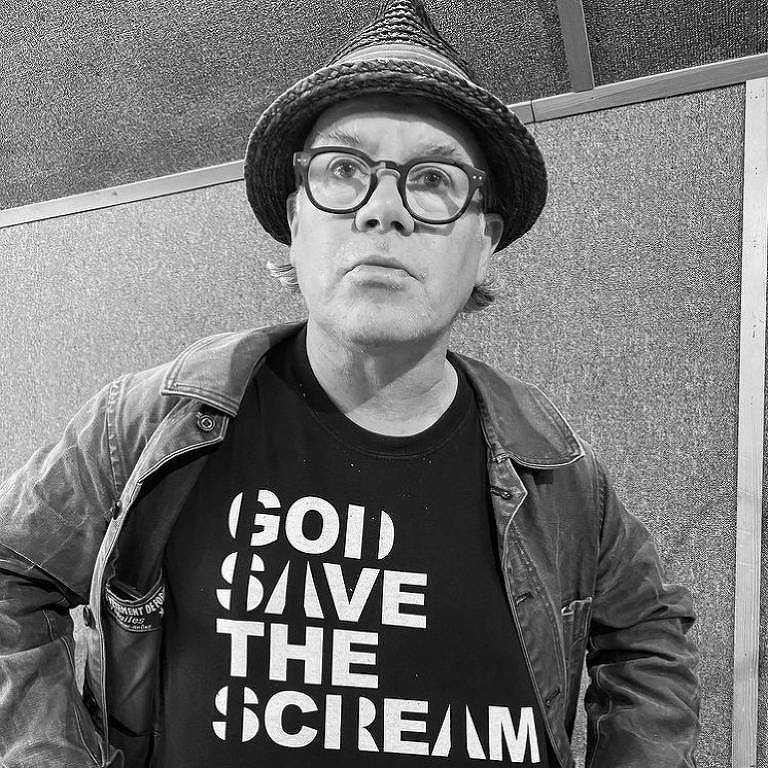 Foto em preto e branco de um homem branco de óculos e chapéu usando uma camiseta que diz 'Deus salve o Scream', em referência à banda Primal Scream