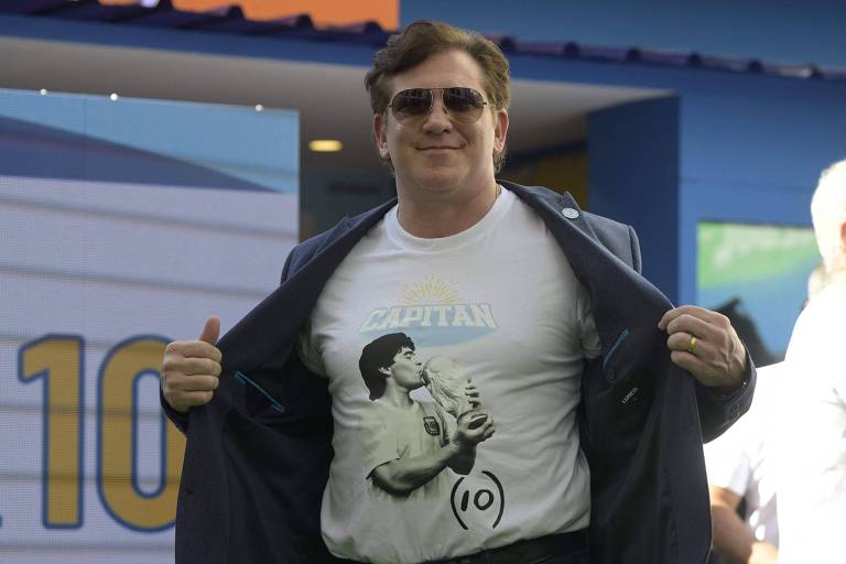 Com óculos escuros e sorrindo, o presidente da Conmebol, Alejandro Domínguez, posa para foto usando camiseta em homenagem a Diego Maradona em cerimônia de homenagem ao segundo ano da morte do craque argentino, em Doha, no Qatar; na foto na camiseta, Maradona geija a Taça Fifa