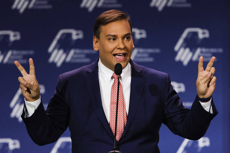 Homem com terno e gravata vermelha fala em frente a um microfone com duas mãos levantadas fazendo sinal de "V"