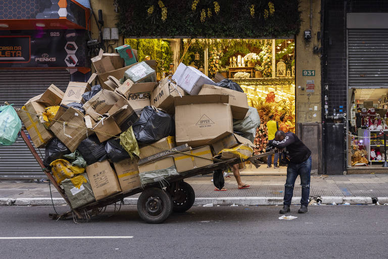 Imagem mostra catador descansando em uma carroça cheia de materias como sacolas plásticas e caixas de papelão. Ele está em uma rua. Ao fundo, há uma calçada com lojas.