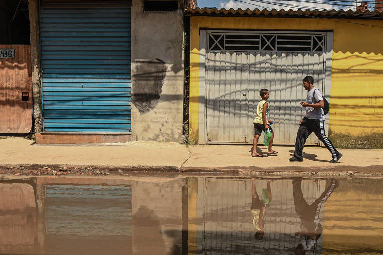 Foto mostra água parada em rua do Jardim Pantanal, na zona leste de São Paulo, e dois jovens andando na calçada