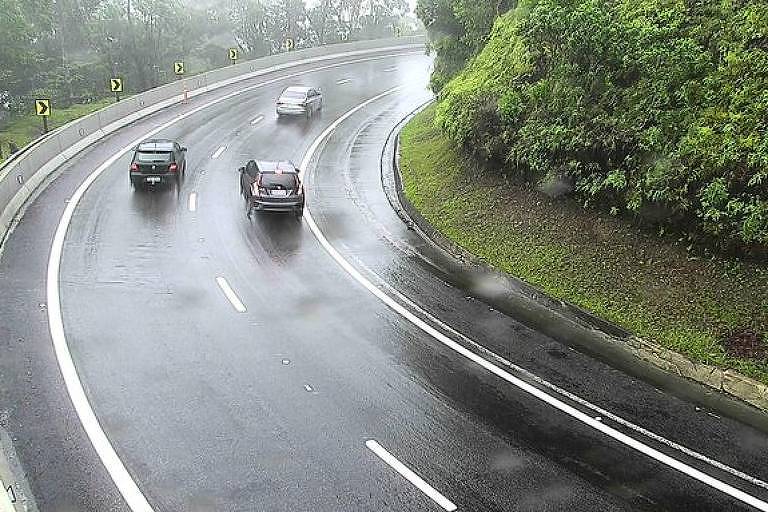 curva de rodovia com três carros, chove, é possível ver mata ao lado direito