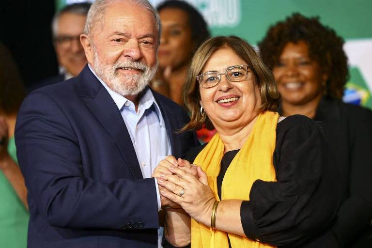 Lula à esquerda, de paletó azul escuro e camisa clara, segura a mão de Cida Gonçalves, que usa óculos e está com uma blusa escura e echarpe amarelo