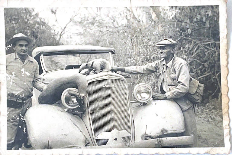 Dois homens posam para foto com animais mortos sobre o carro; um deles segura um pequeno bicho sobre o capô do carro