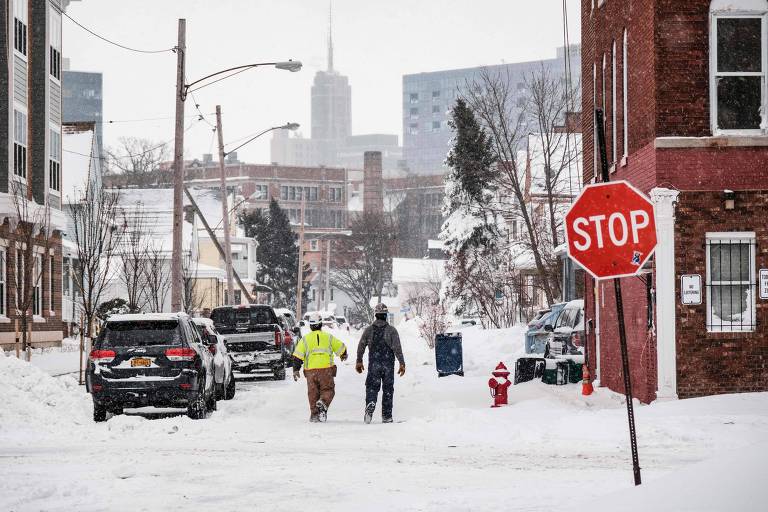 Dois homens, funcionários da companhia de energia elétrica, caminham pela neve acumulada por uma rua enquanto inspecionam as linhas de distribuição



