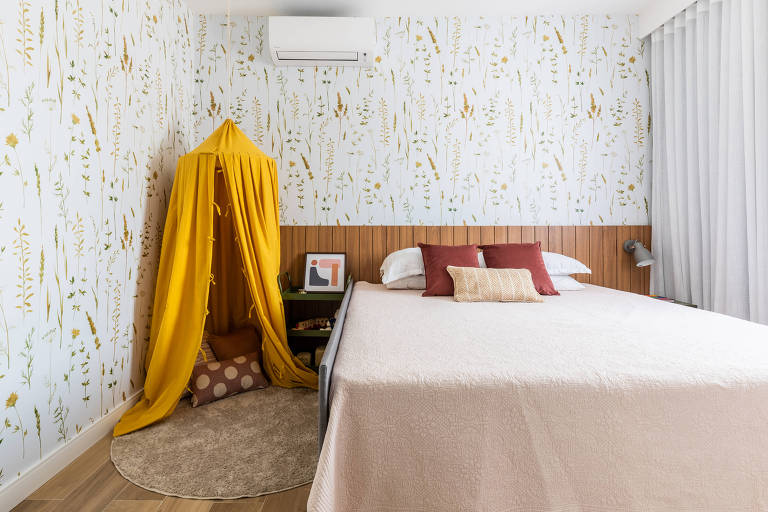 Quarto com cama de casa e, do lado esquerdo, uma tenda amarela pedurada fica em casa de um tapete redondo de pelinho e algumas almofadas