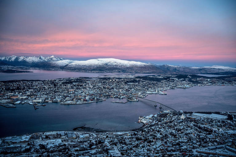 Vista de Tömso, na Noruega, com cidade dividida por um rio; oa fundo é visto um céu arroseado e um cume com neve