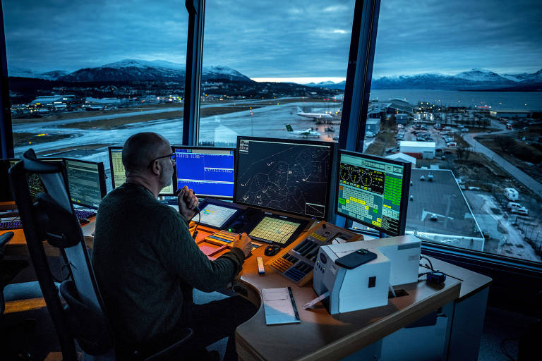Homem sentado monitora telas de computadores; depois da janela de vidro, é visto o aeroporto de Trömso