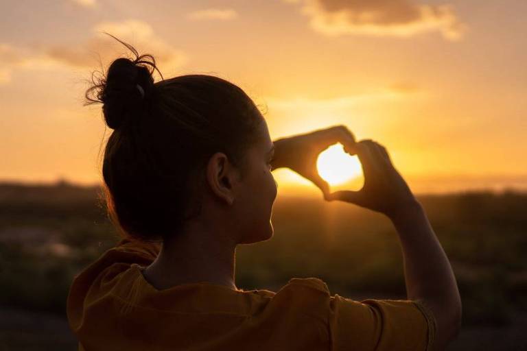 Silhueta contra-luz de mulher, com coque, fazendo sinal de coração com as mãos em direção ao pôr-do-sol