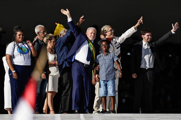Entre várias pessoas que acenam com as mãos para o alto está o presidente Lula, com terno azul escuro e gravata, durante evento da posse 
