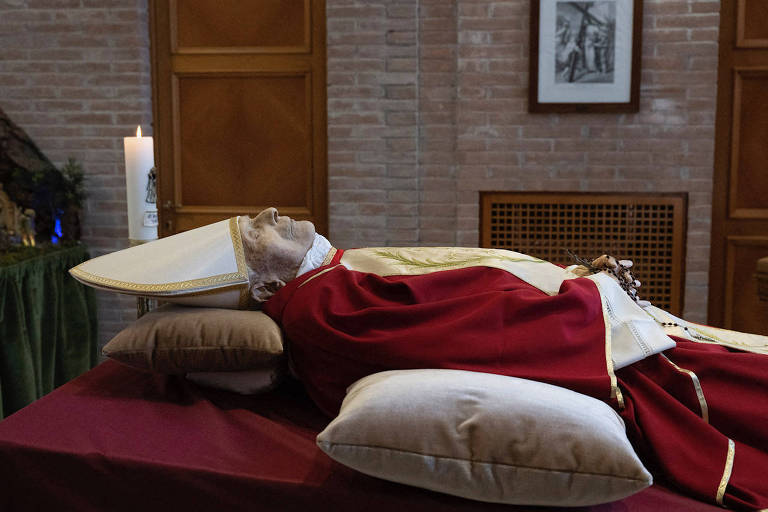 Com vestimentas vermelhas e chapéu pontiagudo branco, o corpo do papa emérito Bento 16 é visto deitado, pela lateral, na capela do mosteiro Mater Ecclesiae, no Vaticano 