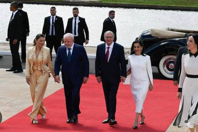 O Presidente Lula e sua mulher, Janja, ela de conjunto dourado, sobem a rampa com Geraldo Alckmin e Dona Lu. Ela usa vestido branco e a seu lado aparece também, fora do tapete vermelho, uma mulher de vestido branco e bolinhas pretas