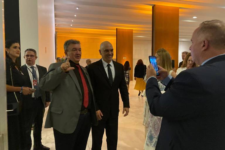 O ministro Alexandre de Moraes, ao lado de um homem que gesticula o L com uma das mãos, num amplo salão. À frente dos dois, uma pessoa registra a cena com um celular 