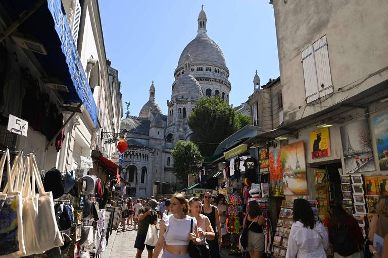 Turistas andam por uma rua de pedrestes iluminada cheia de lojas com mercadorias nas cançadas; ao fundo se vê a cúpula de uma basílica  