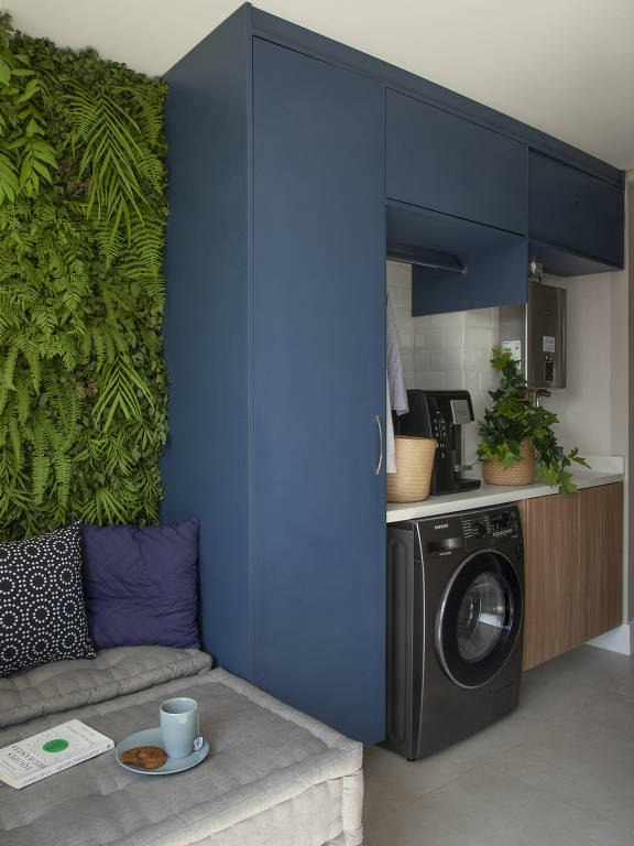 Varanda com painel de plantas e futons. Ao lado, fica a lavanderia com armário azul planejado e máquina de lavar de abertura frontal