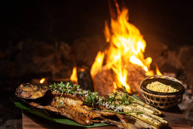 Peixe temperado com pupunha, ao lado uma cumbuca de farofa e as chamas de uma fogueira ao fundo.