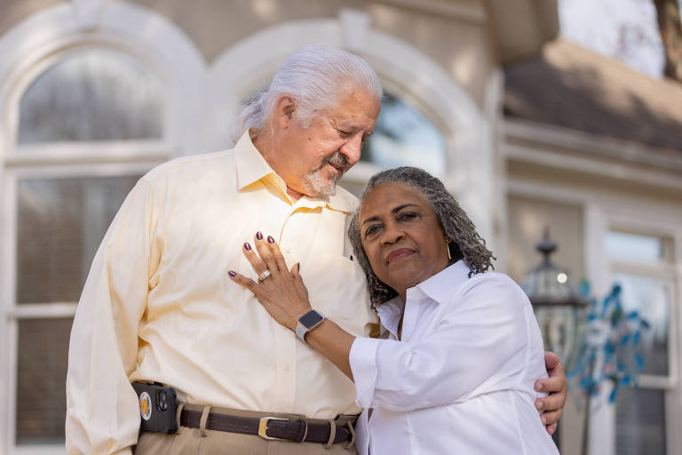 Foto mostra um casal abraçado: à esquerda está um homem de camisa amarelo clara que olha para a mulher; à direita, mulher de camisa brança olha em direção à câmera  