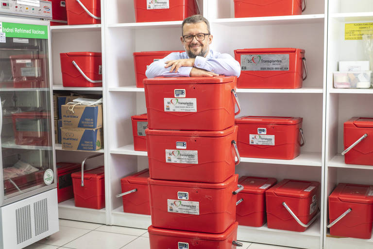 Homem branco, de óculos, veste camisa clara e calça jeans; ele se apoia em três caixas vermelhas que são usadas para transportar órgãos para transplantes