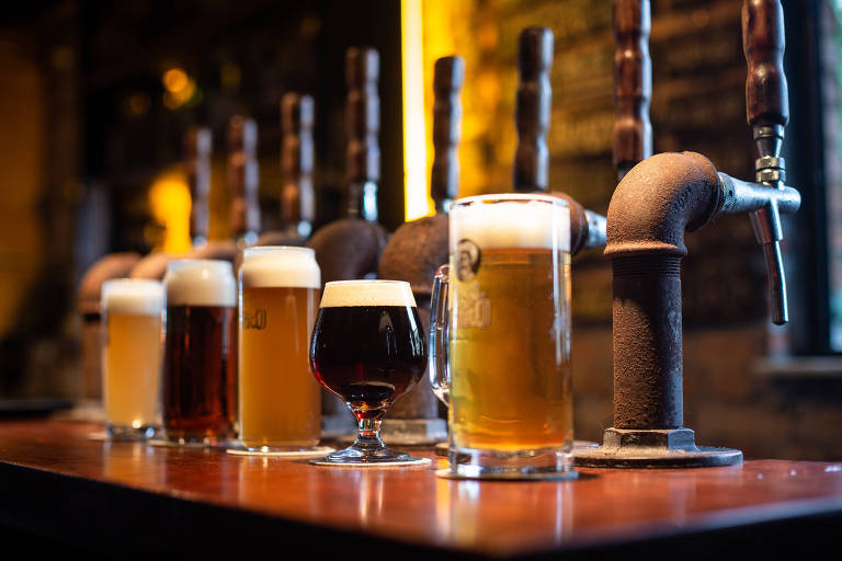 Foto mostra torneiras de chope e cinco copos à frente, cada um correspondente ao tipo de cerveja disponível na torneira