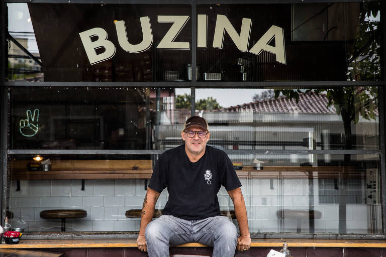 homem branco, de óculos e boné, está sentado em um balcão, onde se lê Buzina ao fundo
