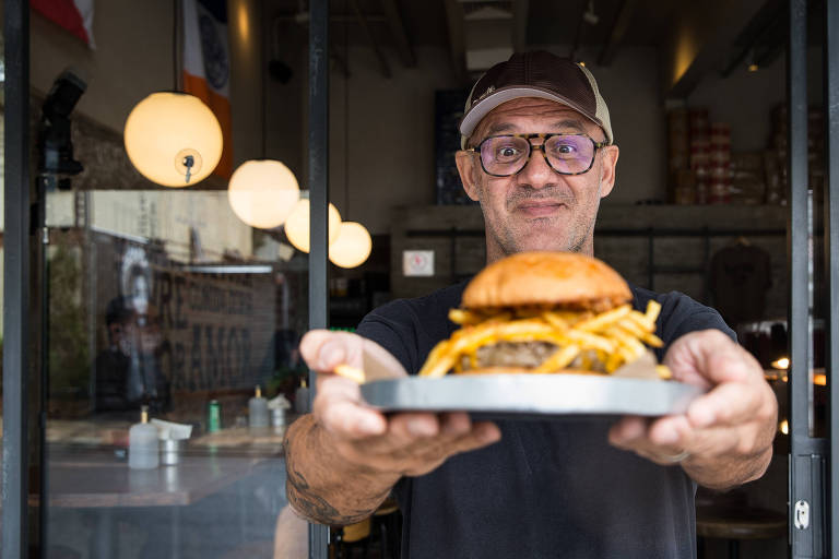 Homem branco, de óculos e boné, segura um hambúrguer com batata frita