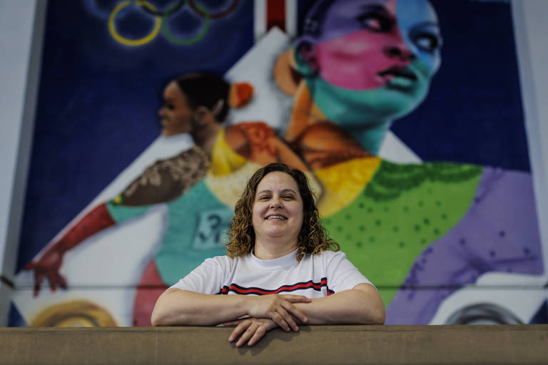 Mulher branca é retratada sorrindo em frente a um painel colorido com o desenho da atleta Rebeca Andrade, no Ginásio Bonifácio Cardoso, em Guarulhos; ela usa camiseta branca com uma faixa vermelha e tem os braços apoiados em uma trave olímpica. 