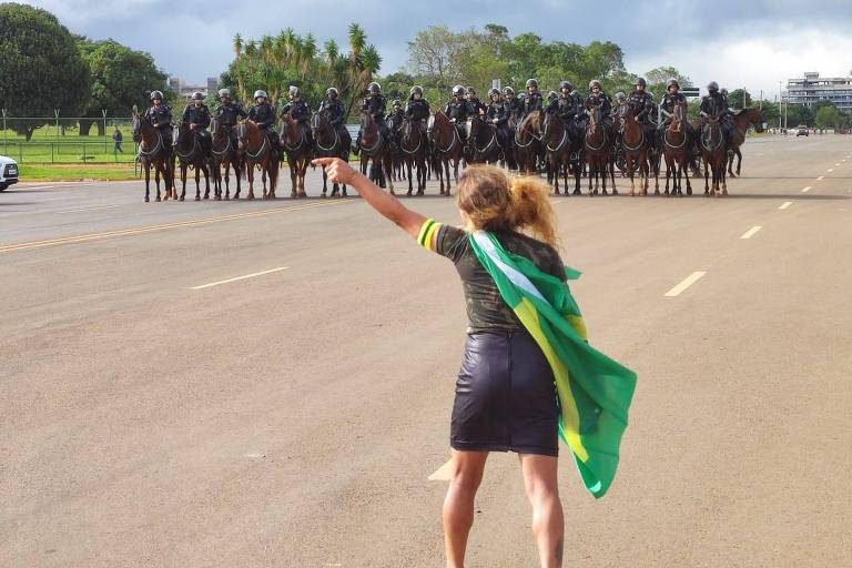Mulher com bandeira do Brasil amarrada junto ao corpo aparece de costas. Diante dela, policiais da cavalaria formam uma barreira na avenida