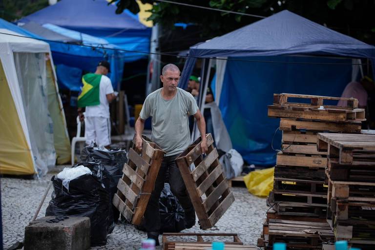 Homem carrega caixotes de madeira em frente a barracas azuis e brancas