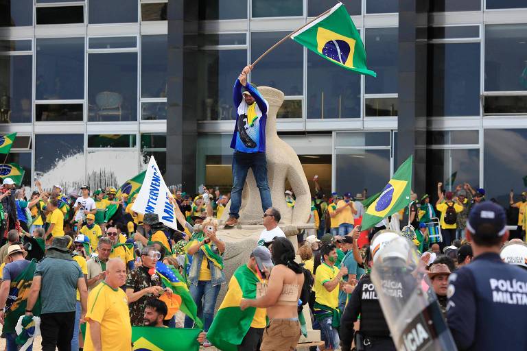 Várias pessoas vestidas de amarelo com bandeiras do Brasil na área externa de um prédio com vidraças. Um manifestante, vestido de azul e jeans, está em pé sobre a estátua da Justiça, agitando uma bandeira do Brasil