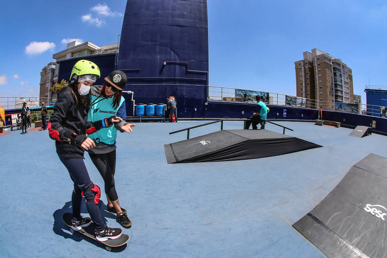 Menina equipada com capacete e tornozeleira em cima de um skate enquanto é segurada por uma mulher